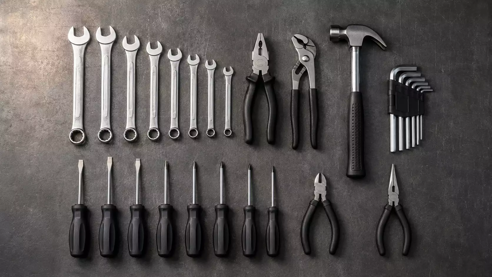 Flat lay of chrome vanadium hand tools including spanners, pliers, hammer, screwdrivers, and allen keys on industrial steel background